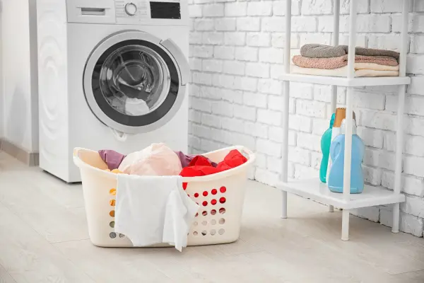 Điểm danh 3 dòng nước giặt cho từng loại máy giặt 2 Một máy giặt, giỏ đựng quần áo bẩn, và kệ chứa các chai nước giặt.