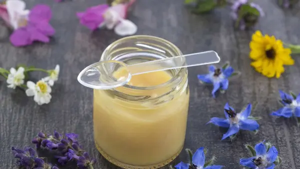 Mẹo bảo quản sữa ong chúa không cần tủ lạnh vẫn giữ trọn dưỡng chất | Điện Máy Gia Phát VN 2 Hũ mật ong mở nắp với thìa nhựa, cùng hoa tươi rải quanh trên bề mặt gỗ.